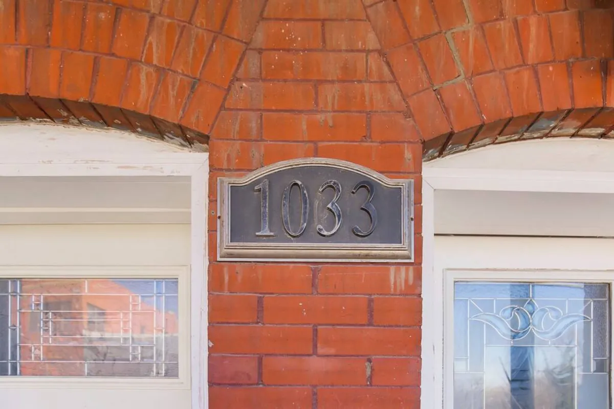A close-up of the house number '1033' under a brick archway at the main entrance, with double front doors. The scene is tidily maintained and projects a sense of security and straightforward access for new arrivals.