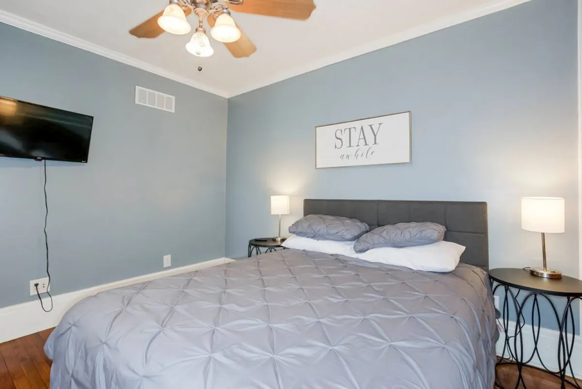 A bright and calming bedroom with a bed flanked by nightstands, soft wall art, and a television. Ample natural light streams in, creating a welcoming and restful environment.