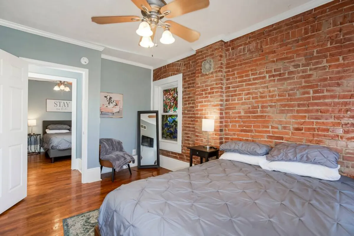 A cozy bedroom scene featuring a bed with plush gray bedding set against an exposed brick wall, flanked by a nightstand with a lamp, a mirror, and tasteful artwork. Soft natural light pours into the room, creating an inviting and restful atmosphere.