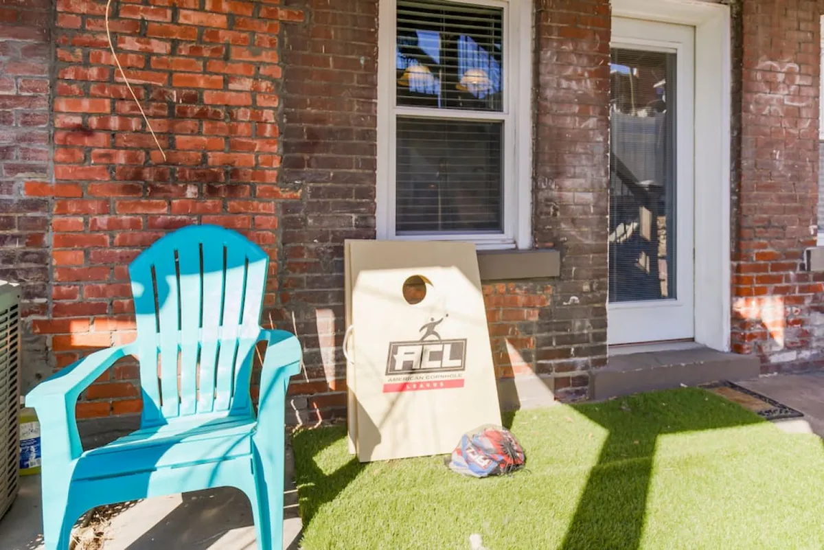 This exterior image highlights a playful patio scene with a turquoise chair, cornhole board, and artificial grass set against a brick wall. The space invites casual outdoor games and friendly gatherings, emphasizing fun and leisure right at the doorstep.