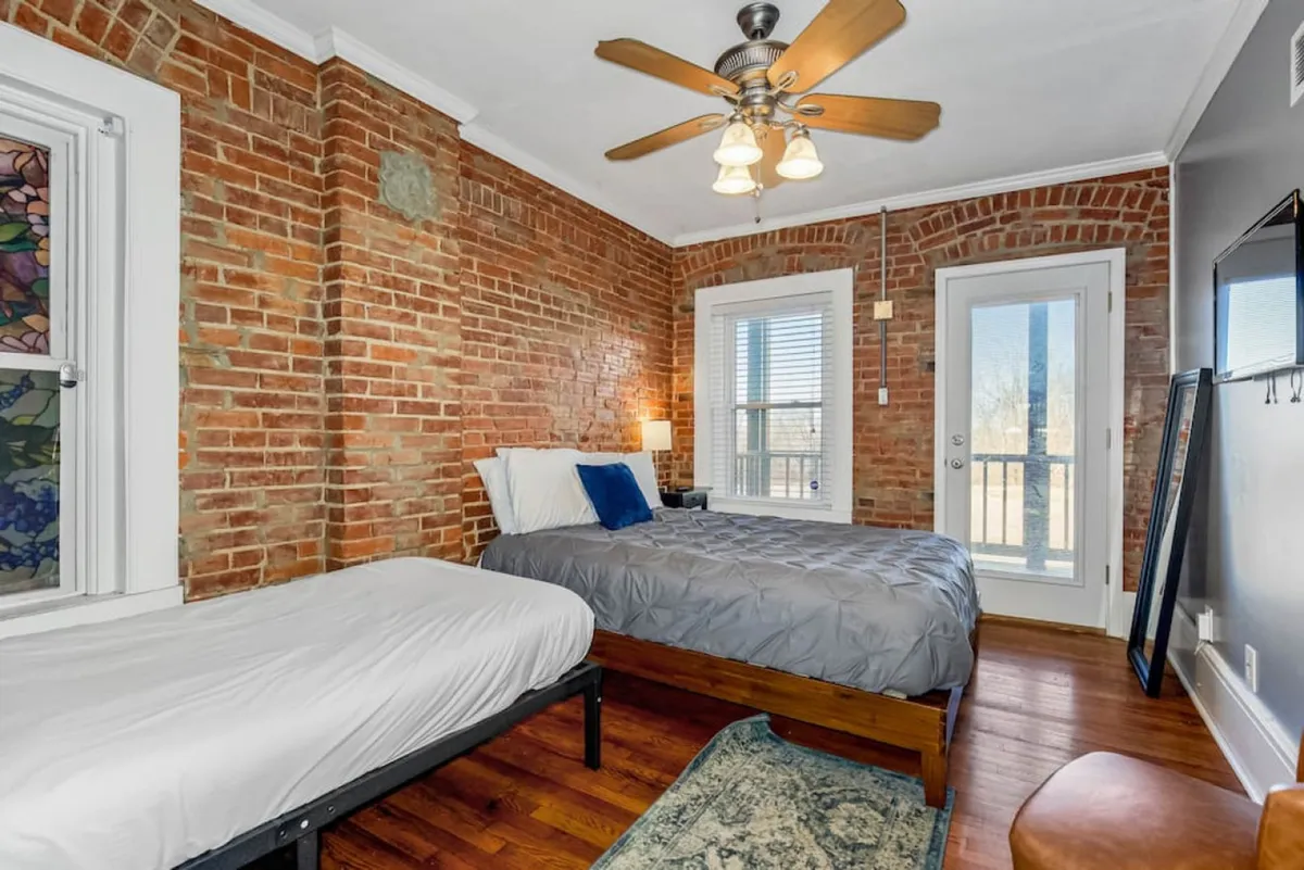 An inviting bedroom featuring exposed brick walls, a plush bed with gray bedding, a stained glass window, and a door leading out to a balcony. The room feels spacious yet intimate, blending warmth and hotel-like luxury.