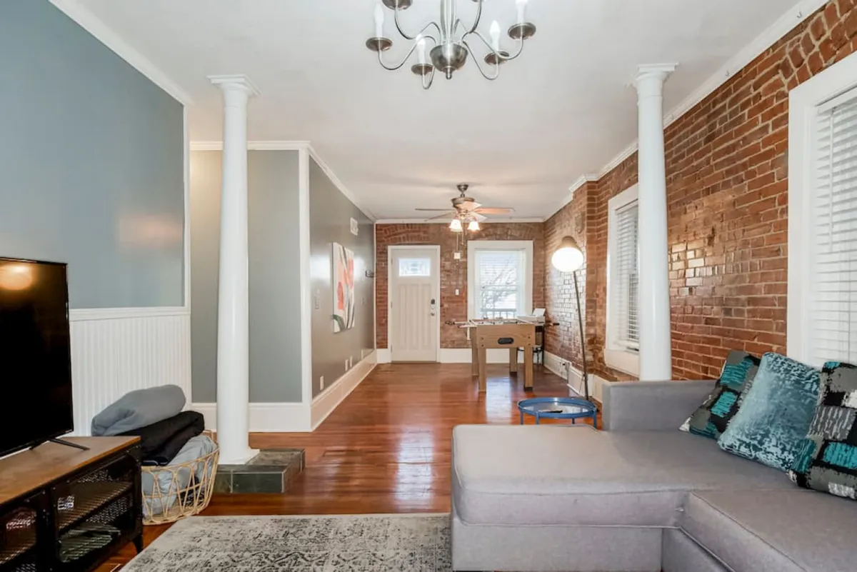 A welcoming open-concept living and dining area with a comfortable sofa, exposed brick wall, elegant chandelier, and a dining table set for gatherings. The space is designed for both relaxing with friends and entertaining, with a stylish yet homey ambiance.
