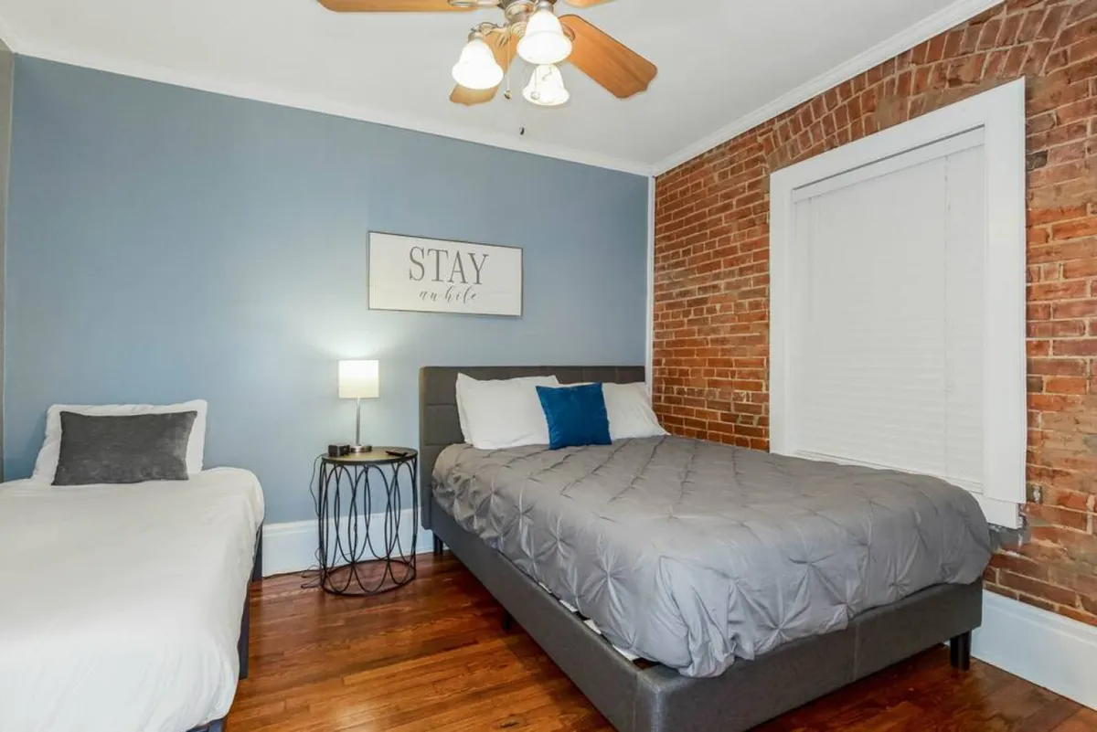 A tranquil interior corner with a comfortable bed, exposed brick wall, framed sign, and warm lamp lighting. The scene feels welcoming and peaceful—ideal for unwinding after a busy day.