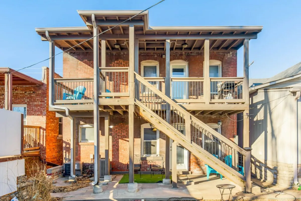 The exterior of a brick building with a welcoming wooden staircase, balconies adorned with blue chairs, and a view that suggests relaxing outdoor moments. The relaxed setting hints at social gatherings or peaceful solo mornings, all framed by the lively brick facade.