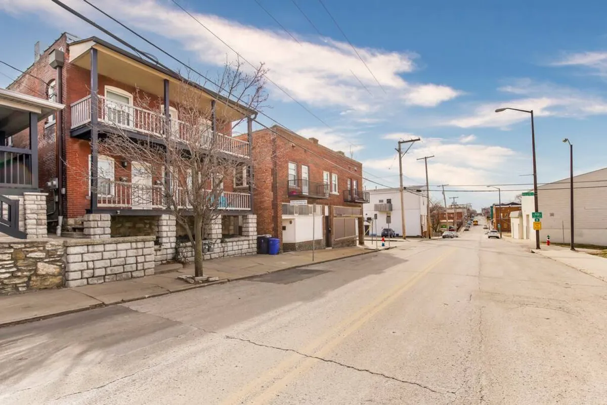 A sunlit neighborhood street lined with classic brick buildings, leafy street trees, power lines overhead, and a wide sidewalk inviting people to walk, explore, and mingle. The urban environment captures the essence of community and accessibility, hinting at nearby shops and local attractions just steps away.