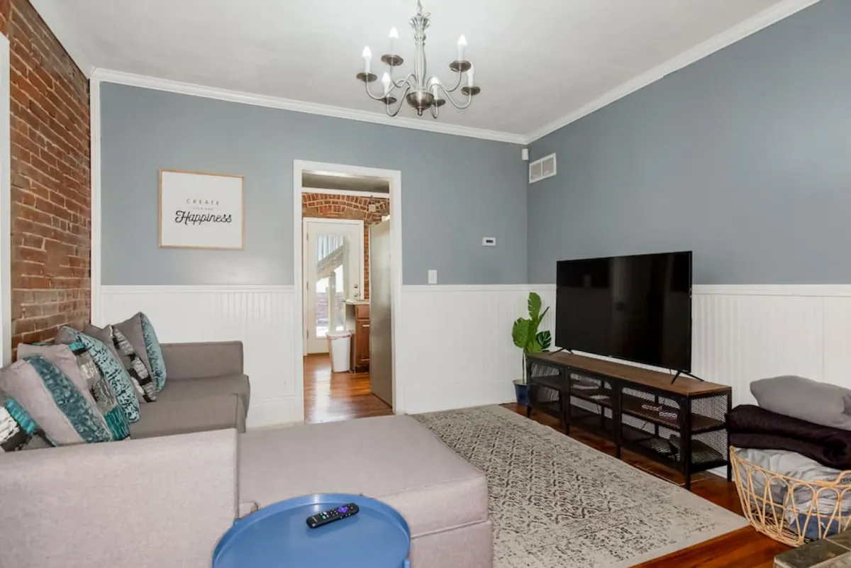 The image shows a cozy living area with a plush sofa adorned with colorful cushions, a modern television stand, a striking chandelier, and an exposed brick wall that adds urban charm. The space is designed for comfort, perfect for lounging, watching TV, or gathering with friends and family.