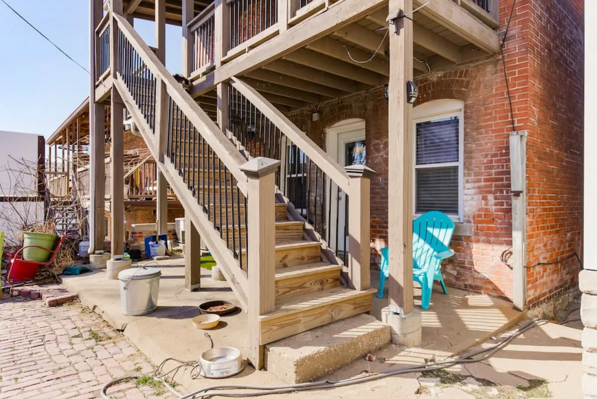 This exterior image features a warmly lit wooden staircase against a classic brick wall, with a blue outdoor chair invitingly positioned on the landing. The scene captures the ambiance of relaxing outdoors in a city setting, suggesting a peaceful spot to unwind after exploring urban attractions. Hints of sunset add to the cozy, welcoming atmosphere.