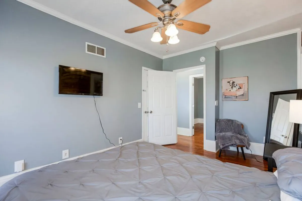 This image showcases a private bedroom with a plush bed, wall-mounted television, stylish artwork, a mirror, and a comfortable chair in the corner. The room exudes a tranquil atmosphere, ideal for relaxation and unwinding.