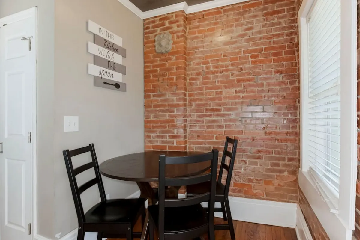 A cozy dining area with a round table, black chairs, exposed brick wall, and a decorative wall sign. The setting is intimate and inviting, designed for casual dining, morning coffee, or small gatherings, encouraging conversation and shared meals.