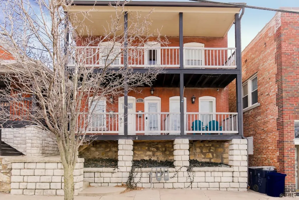 This image shows a welcoming exterior balcony with comfortable chairs, an exposed brick facade, and a decorative stone base. The balcony overlooks the neighborhood, suggesting a connection to the local area and a place to relax before or after attending nearby events.