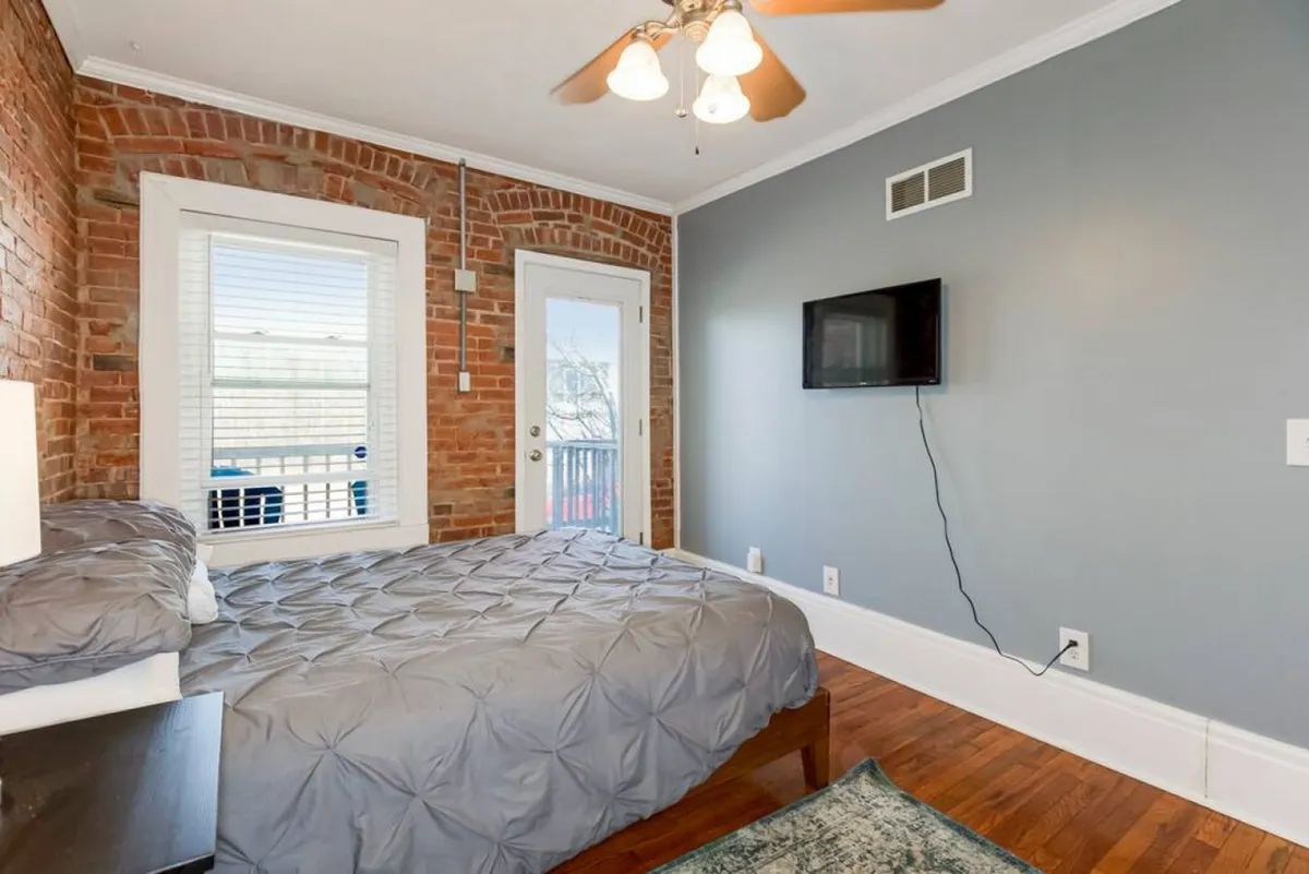 A view of a comfortable bed set against an exposed brick wall, with a door leading to a private balcony and a mounted television. The space is bright and inviting, balancing relaxation with modern convenience.