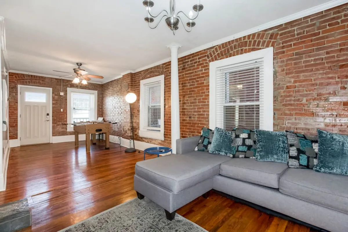 A cozy living area featuring a plush sofa, exposed brick wall, glimmering chandelier, and a game table in the background. The scene is set for intimate gatherings with friends, game nights, or simply relaxing in a stylish, downtown-inspired environment.